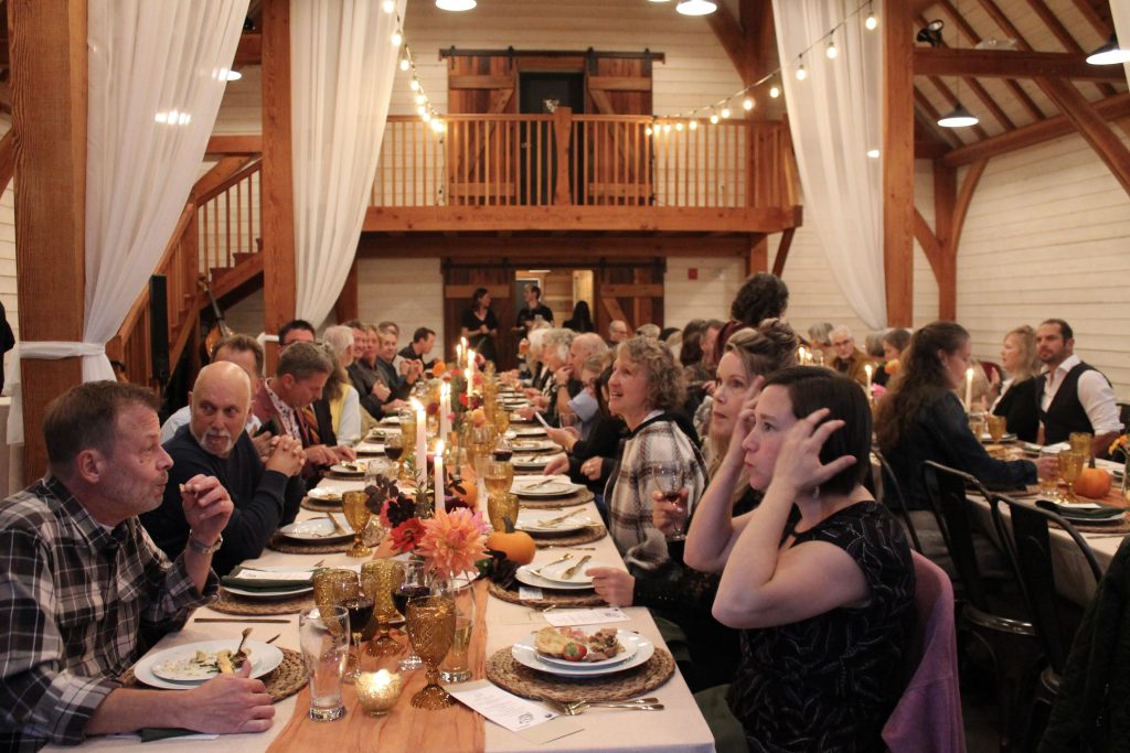 A large group of people sit on either side of a very long dinner table, talking to each other. The table is decorated with flowers and candles and appears to be inside a wooden barn-like building.