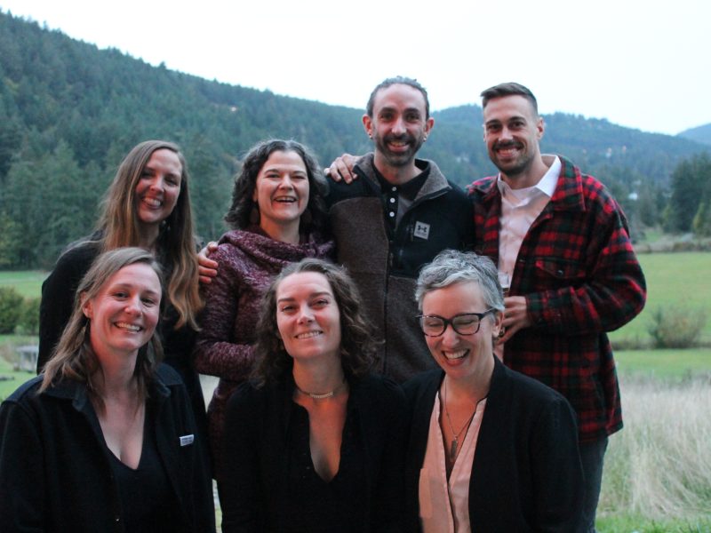 A group of seven people, four standing and three kneeling in front, pose outdoors for a photo with mountains seen behind. They are current and former Cow-op staff.