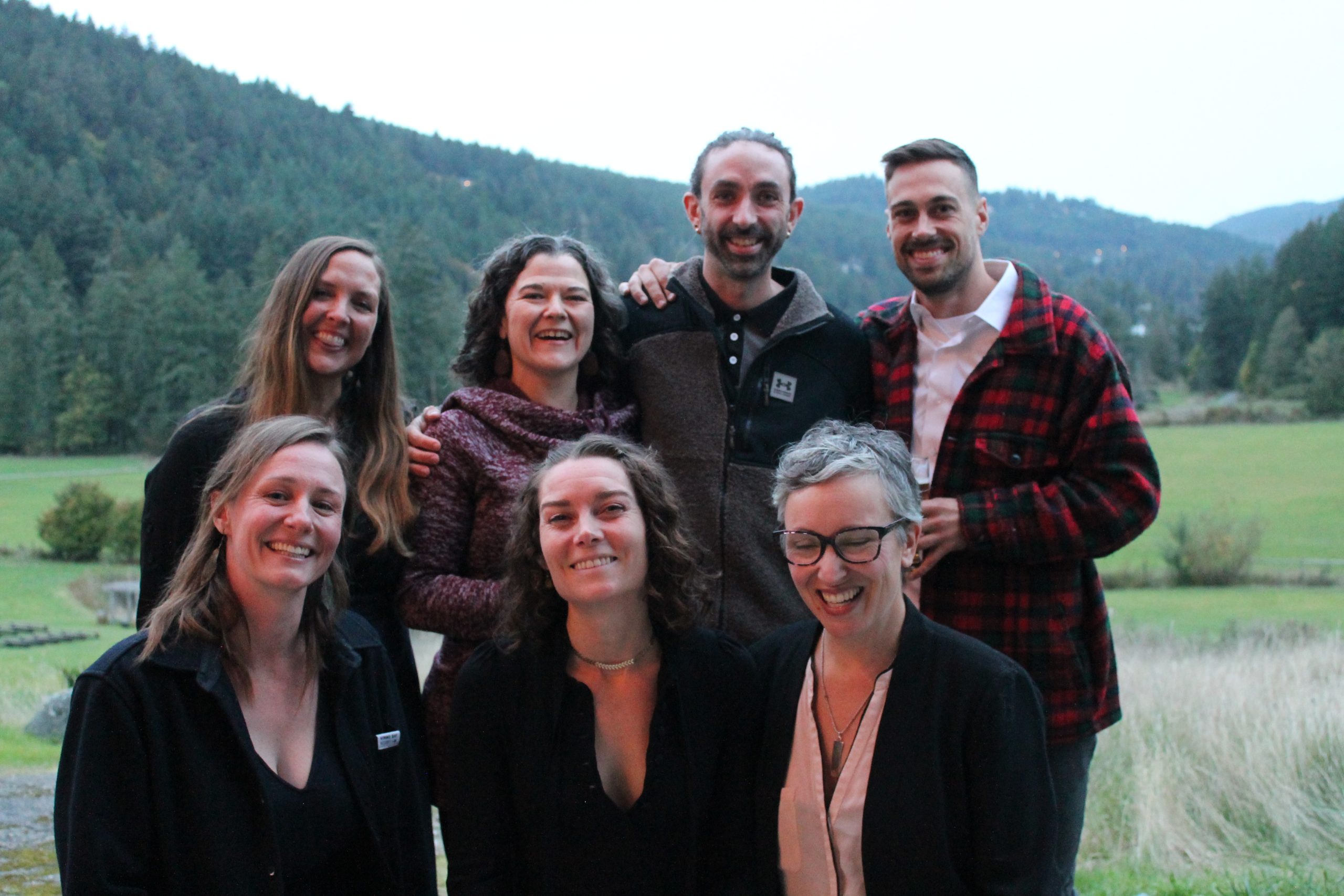A group of seven people, four standing and three kneeling in front, pose outdoors for a photo with mountains seen behind. They are current and former Cow-op staff.