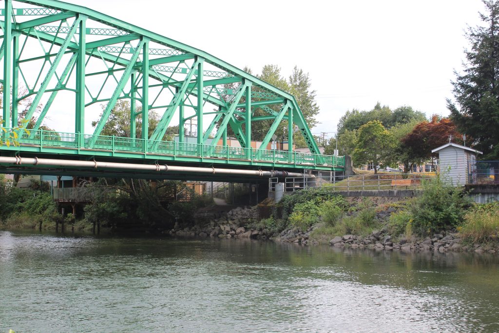 A green bridge with a river below. There are stacked rock on the banks for erosion control.