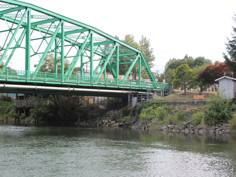 A green bridge with a river below. There are stacked rock on the banks for erosion control.