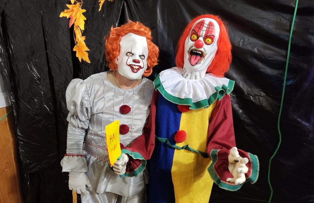 A person dressed in a Pennywise costume stands next to an animatronic clown in front of a black backdrop at the Glenora Haunted Hall