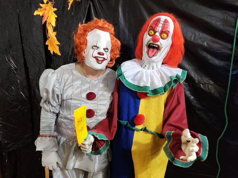 A person dressed in a Pennywise costume stands next to an animatronic clown in front of a black backdrop at the Glenora Haunted Hall