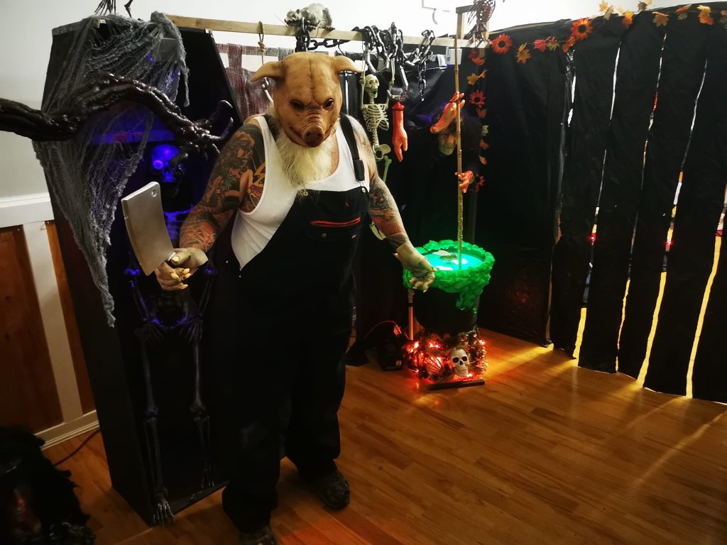 A person in a pig costume holding a cleaver stares at the camera in front of a backdrop of animatronic props.