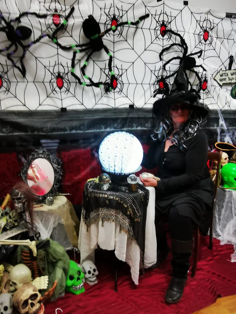 A woman in a witch costume sits at a table with a large light up crystal ball and a backdrop of spiderwebs behind.
