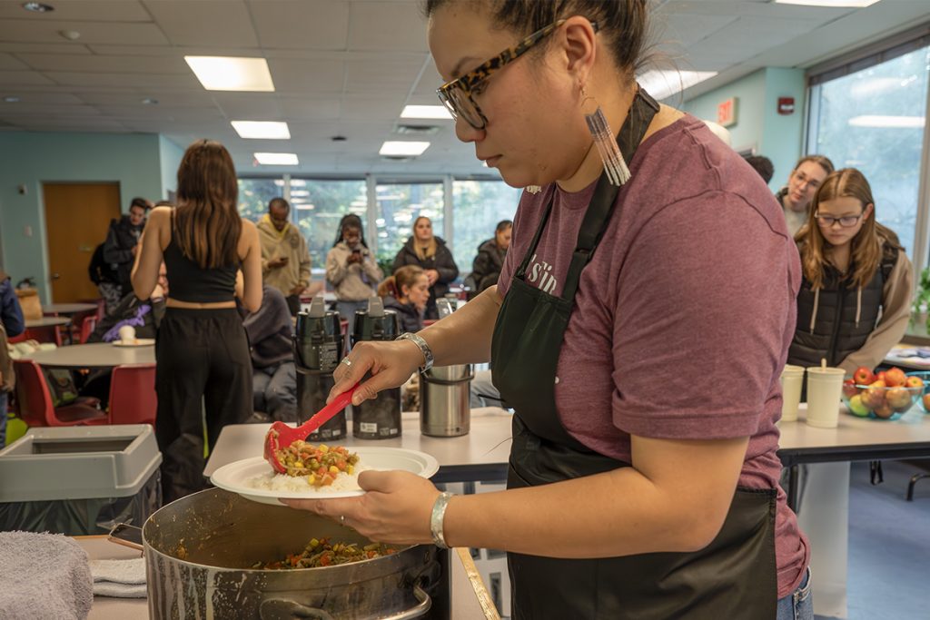 Photo of a woman wearing an apron serving a meal of vegetable curry on rice out of a pot.