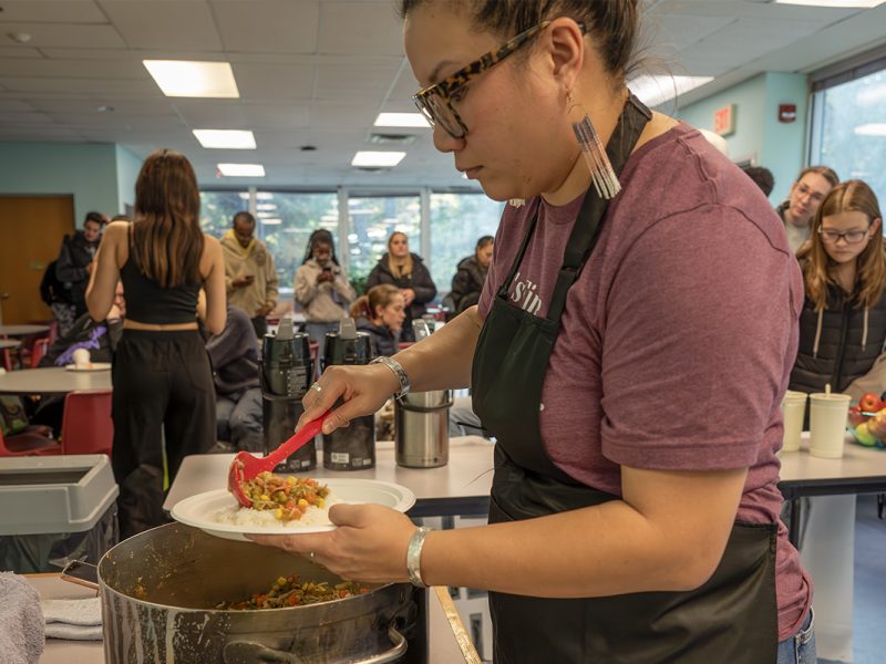 Photo of a woman wearing an apron serving a meal of vegetable curry on rice out of a pot.