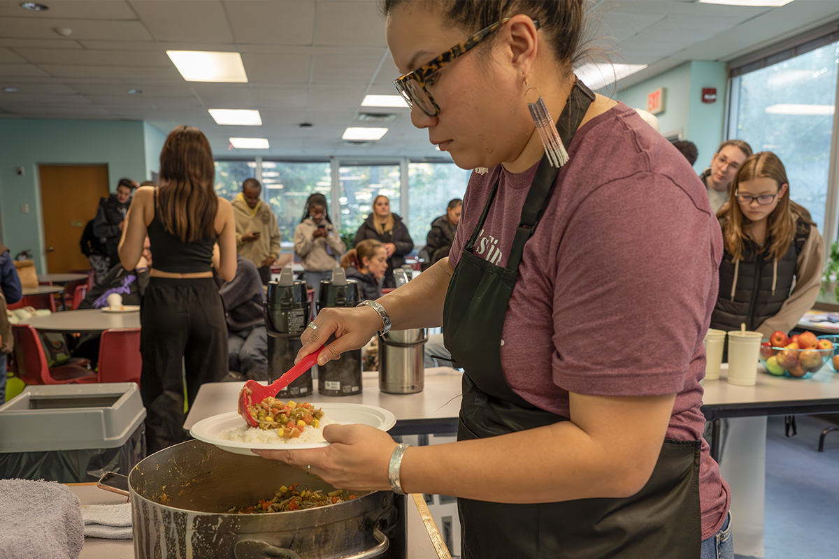Photo of a woman wearing an apron serving a meal of vegetable curry on rice out of a pot.