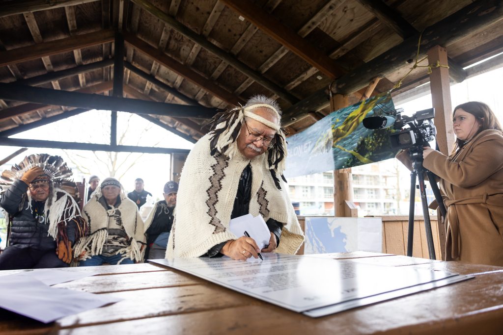 A chief is shown signing a document in traditional regalia.