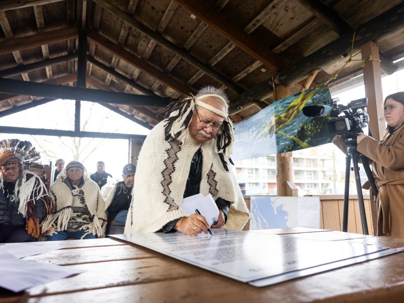 A chief is shown signing a document in traditional regalia.