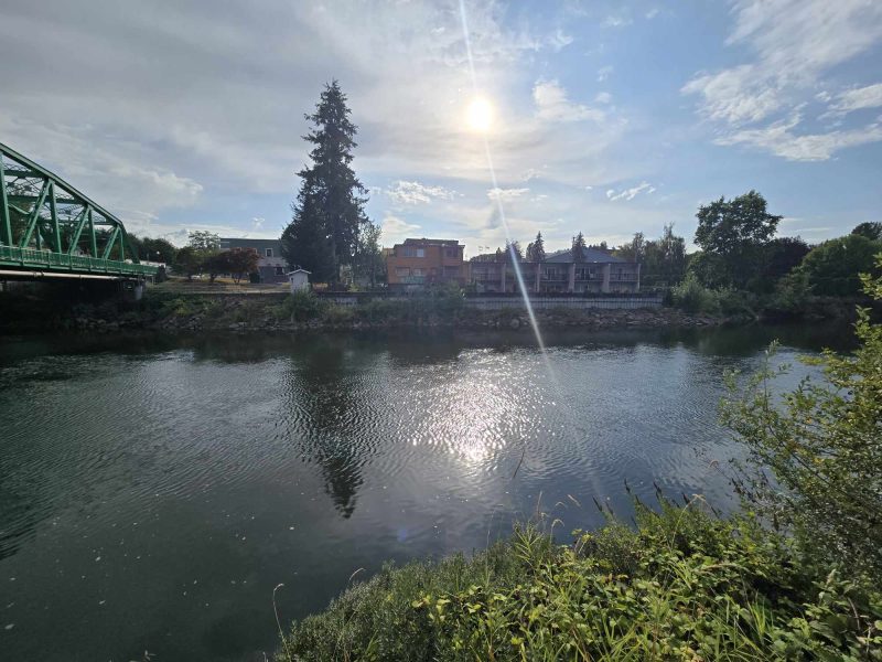 A serene river with calm water reflecting the sunlight. A green metal bridge spans the river on the left, while trees and buildings line the opposite bank. The sky is partially cloudy, and the sun shines high, casting a glow over the landscape.