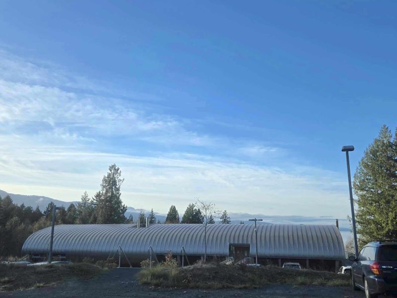 A morning shot of the VIU Deep Bay Marine Research Centre, which has a uniquely-shaped rounded, long metal roof. It looks to be the morning, and it is sunny out.