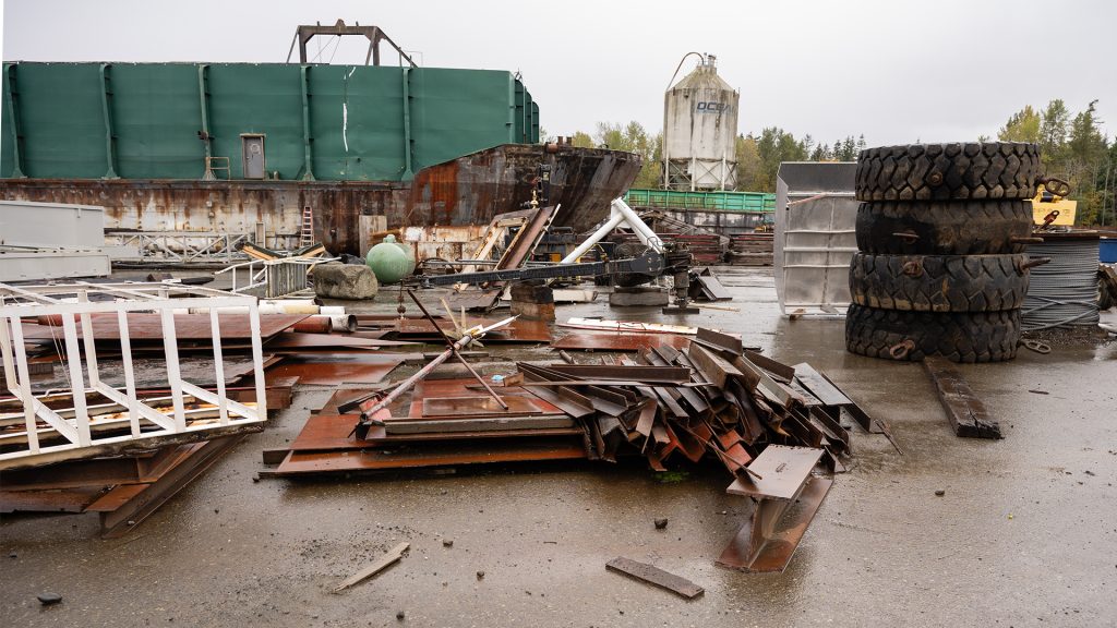 A large barge sits on a beach. It is very rainy. Scrap metal is piled up in front of it, and large tires are piled up to the right.