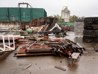 A large barge sits on a beach. It is very rainy. Scrap metal is piled up in front of it, and large tires are piled up to the right.