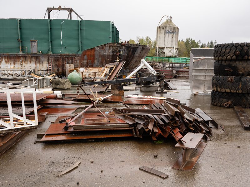 A large barge sits on a beach. It is very rainy. Scrap metal is piled up in front of it, and large tires are piled up to the right.