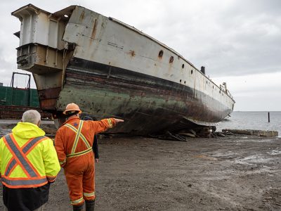 Two men in high visibility outfits stand facing away from the camera. One points at a large ship to his left.