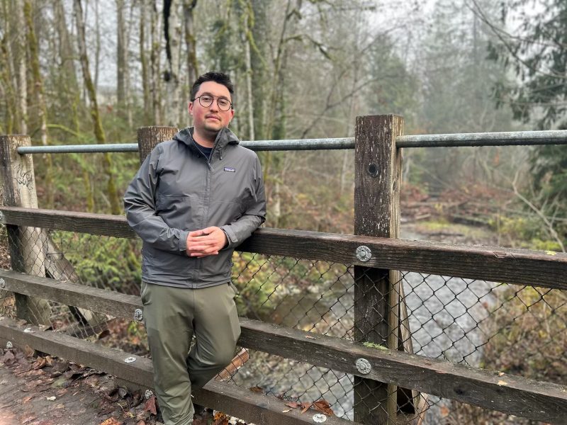 Eric Richards leans against a fence in the forest by a river.