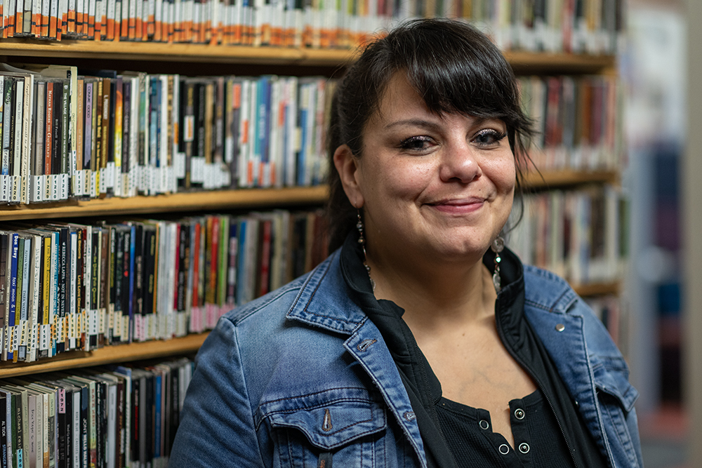 Beverley Planes, an advocate for people who use drugs and overdose prevention, wearing a jean jacket stands in front of a wall of CDs.