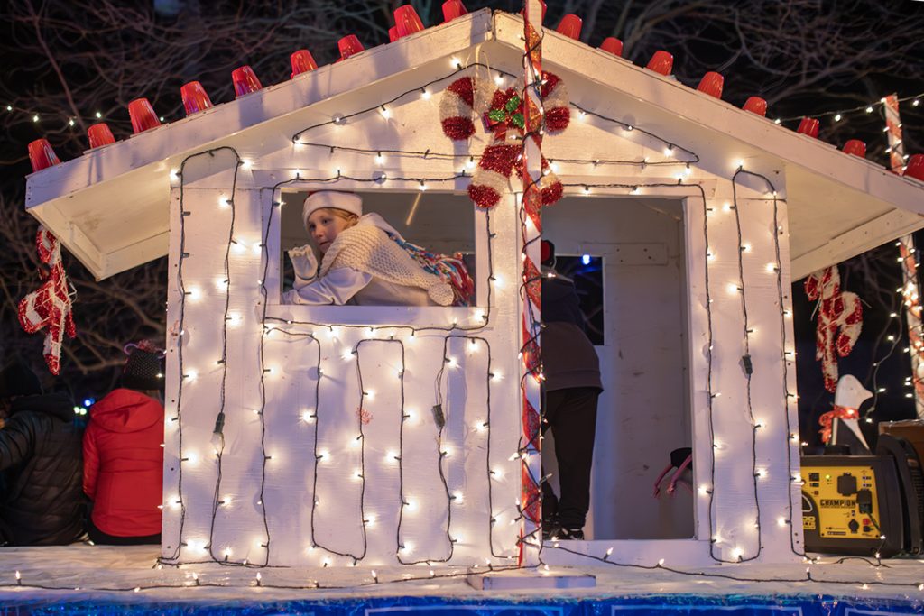 A girl looking out the window of a small wooden shack decorated with Christmas lights waves.