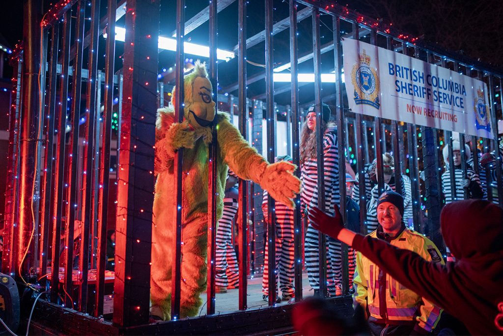 Photo of a person in a Grinch costume and families dressed in prison stripped uniforms in a mock jail float with the words British Columbia Sheriff Service.