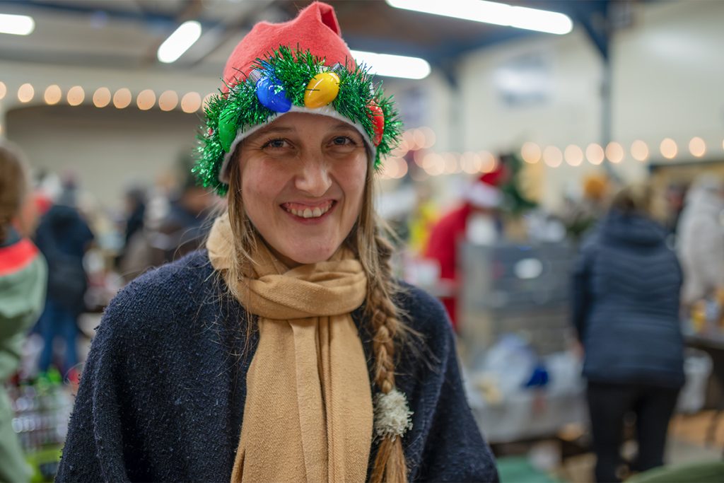 A woman smiling and wearing a Santa hat with lightbulbs and garland trim.