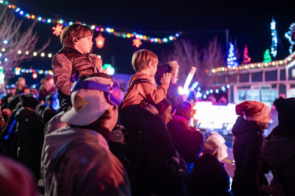 Children sit on top of their parent's shoulders to watch a parade of Christmas lights at night.