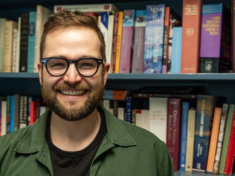 Photo of a bearded man with glasses standing in front of a bookshelf smiling.