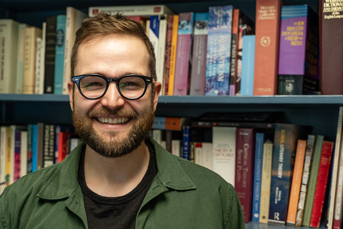 Photo of a bearded man with glasses standing in front of a bookshelf smiling.