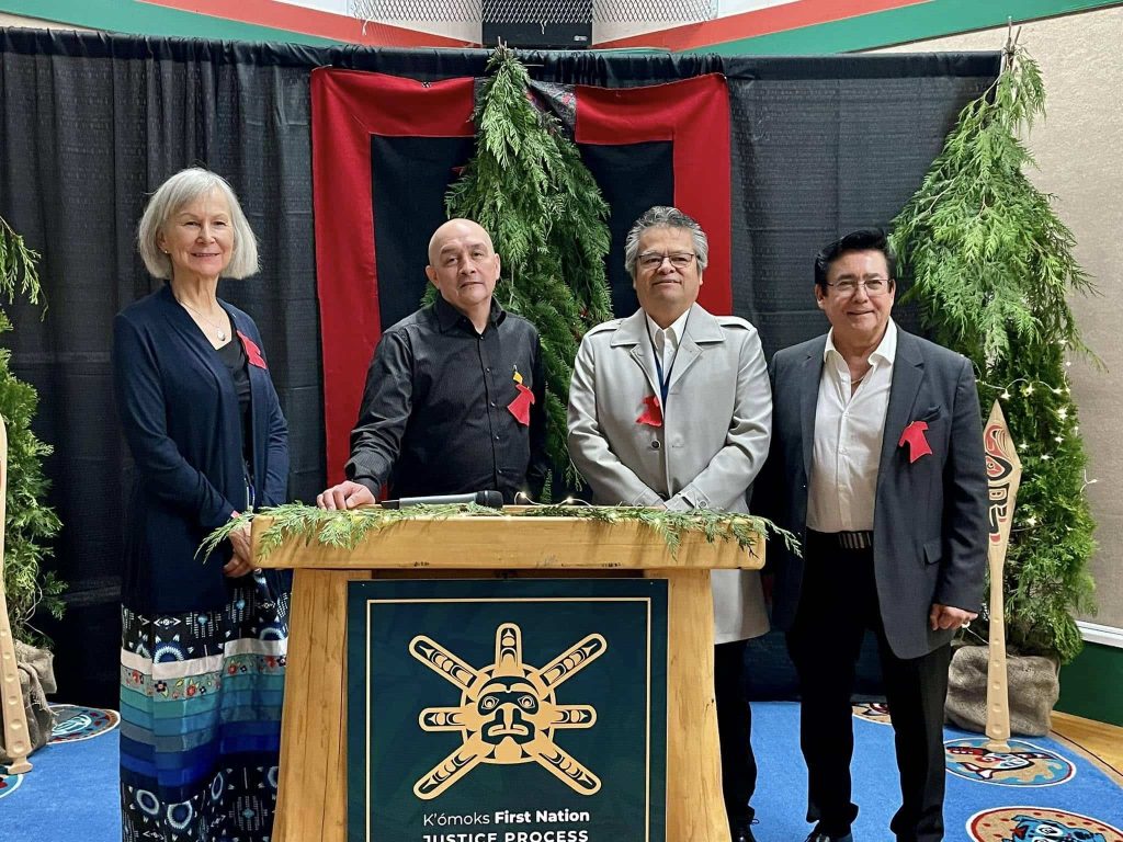 Marion Buller stands to the left of three others behind a podium. There are cedar boughs behind them and the K’ómoks First Nation logo on the podium.