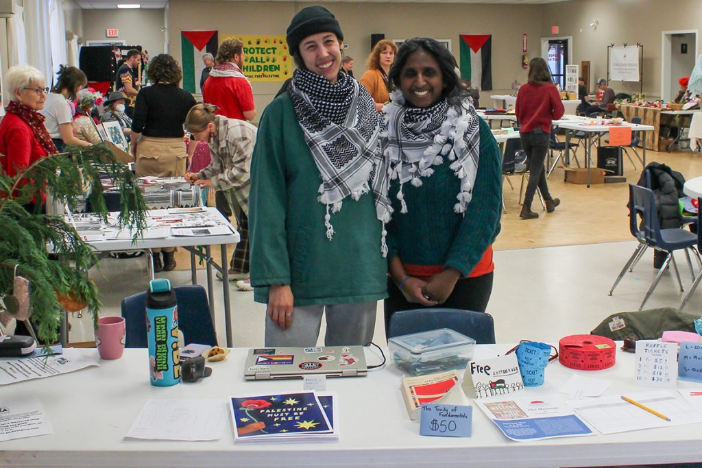 Two women wear Palestinian Keffiyehs and stand in front of a table with various posters and items.