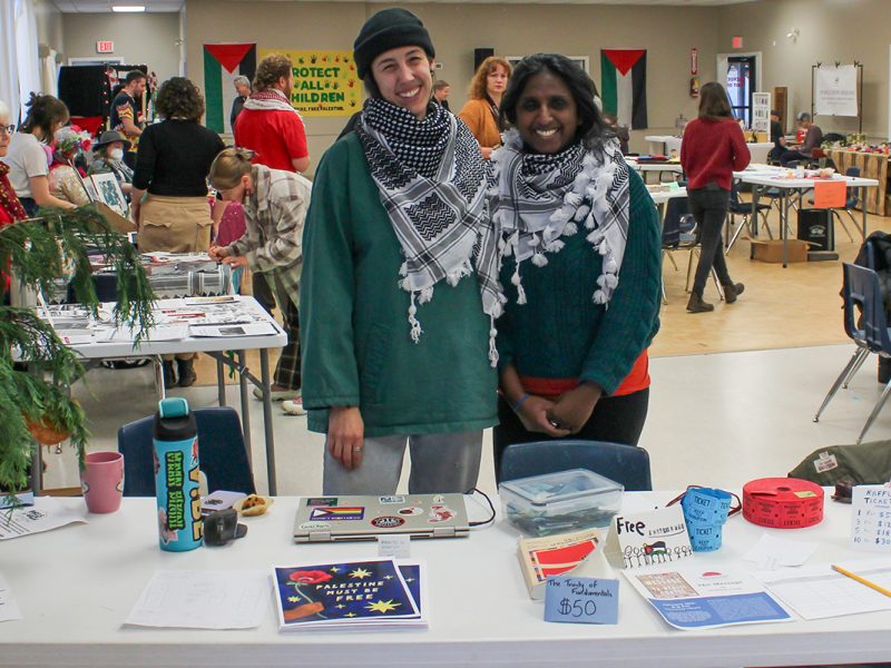 Two women wear palestinian keffiyehs and stand in front of a table with various posters and items