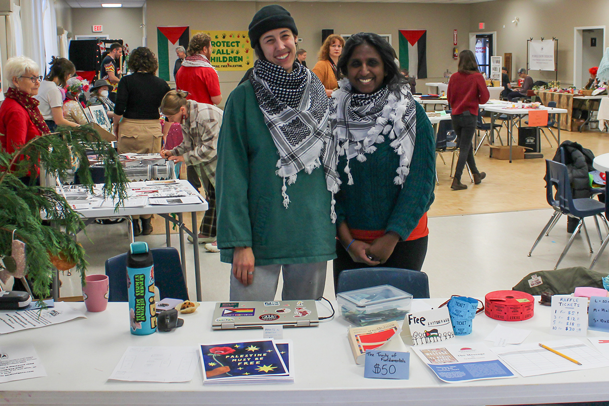 Two women wear palestinian keffiyehs and stand in front of a table with various posters and items