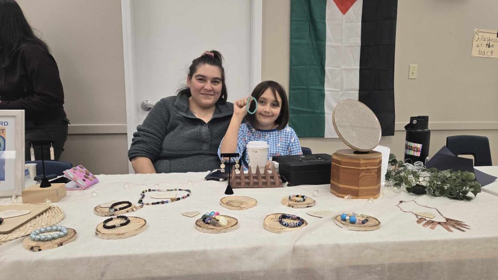 A woman and her child sit next to each other in front of beaded bracelets for sale at the Comox Valley for Palestine winter art market.
