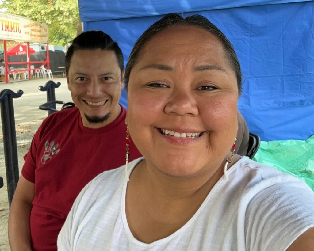 A selfie, taken by Larissa with Clayton behind and to the left. They're both smiling. They are artists featured in the Indigenous Makers Market.