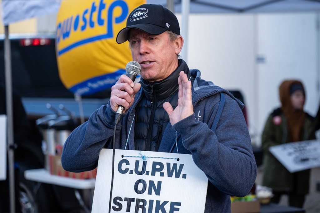 Photo of a man speaking into a microphone while wearing a sign reading C.U.P.W. on strike.