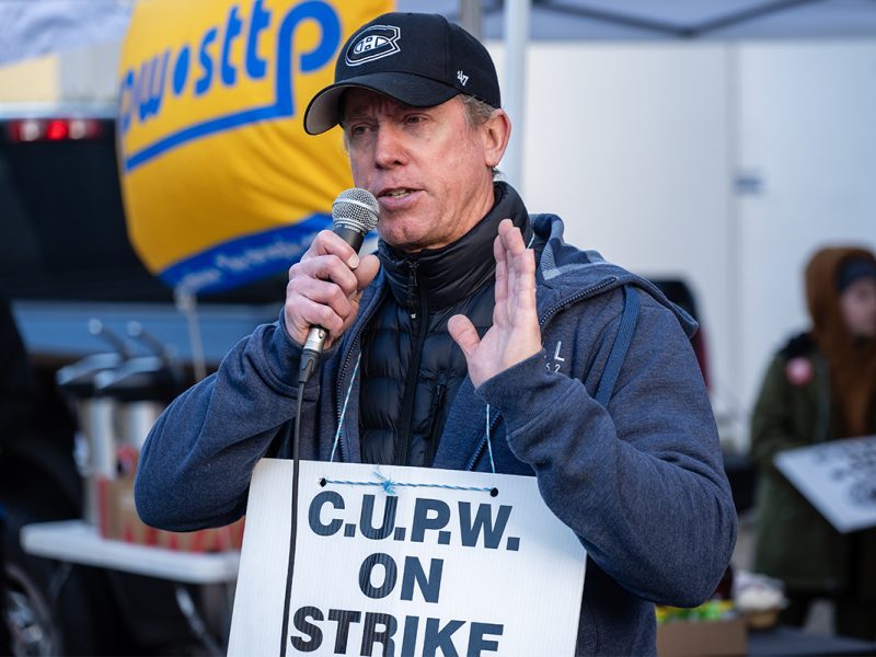 Photo of a man speaking into a microphone while wearing a sign reading C.U.P.W. on strike.