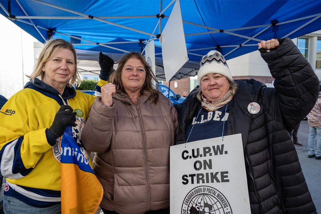 Three women, one of whom is wearing a CUPW on strike sign, pose for a photo with their fists in the air.