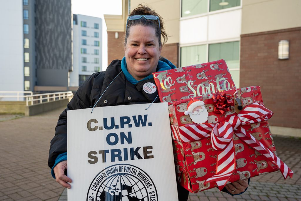 A woman wearing a sign reading C.U.P.W on strike holds a box decorated in wrapping paper and a bow with the words To Santa.