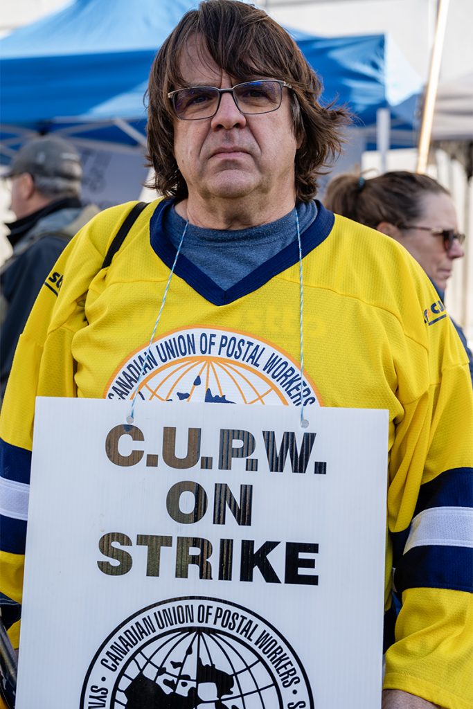 A mean wearing a yellow hockey jersey with the words Canadian Union of Postal Workers wears a sign reading CUPW on Strike.