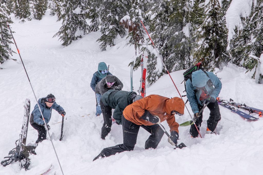 Six people in snow gear dig with small shovels into deep snow during a companion rescue course with Island Alpine Guides in December 2024. The snow has snow probes sticking out of it near where they are digging. Their skis and snowboards have been taken off and sit near them in the snow.