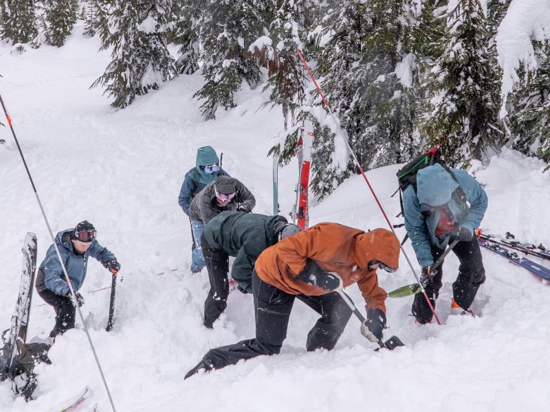 Six people in snow gear dig with small shovels into deep snow during a companion rescue course with Island Alpine Guides in December 2024. The snow has snow probes sticking out of it near where they are digging. Their skis and snowboards have been taken off and sit near them in the snow.