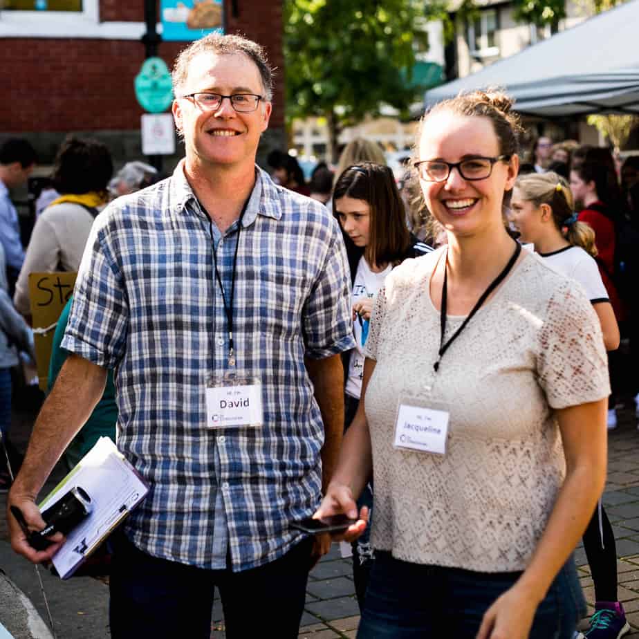 David Minkow and Jacqueline Ronson stand in downtown Duncan with Discourse nametags and notebooks/recorders in hand. They're smiling and facing the camera.