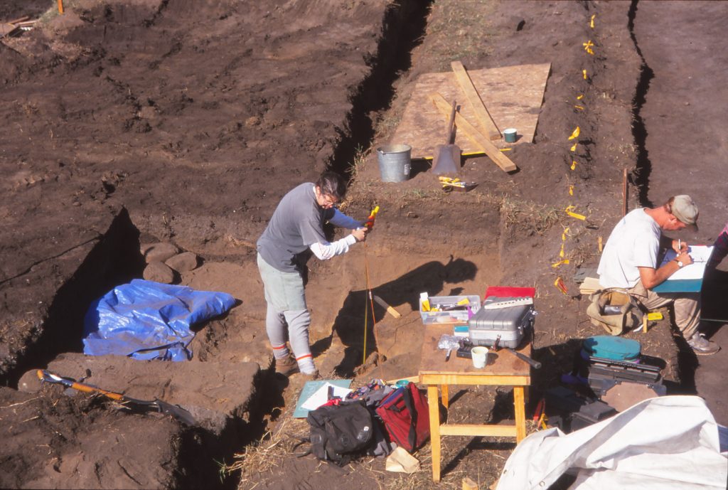 A photo copied from a 1994 slide shows archaeologists looking over tables with equipment surrounded by dirt at an excavation site.