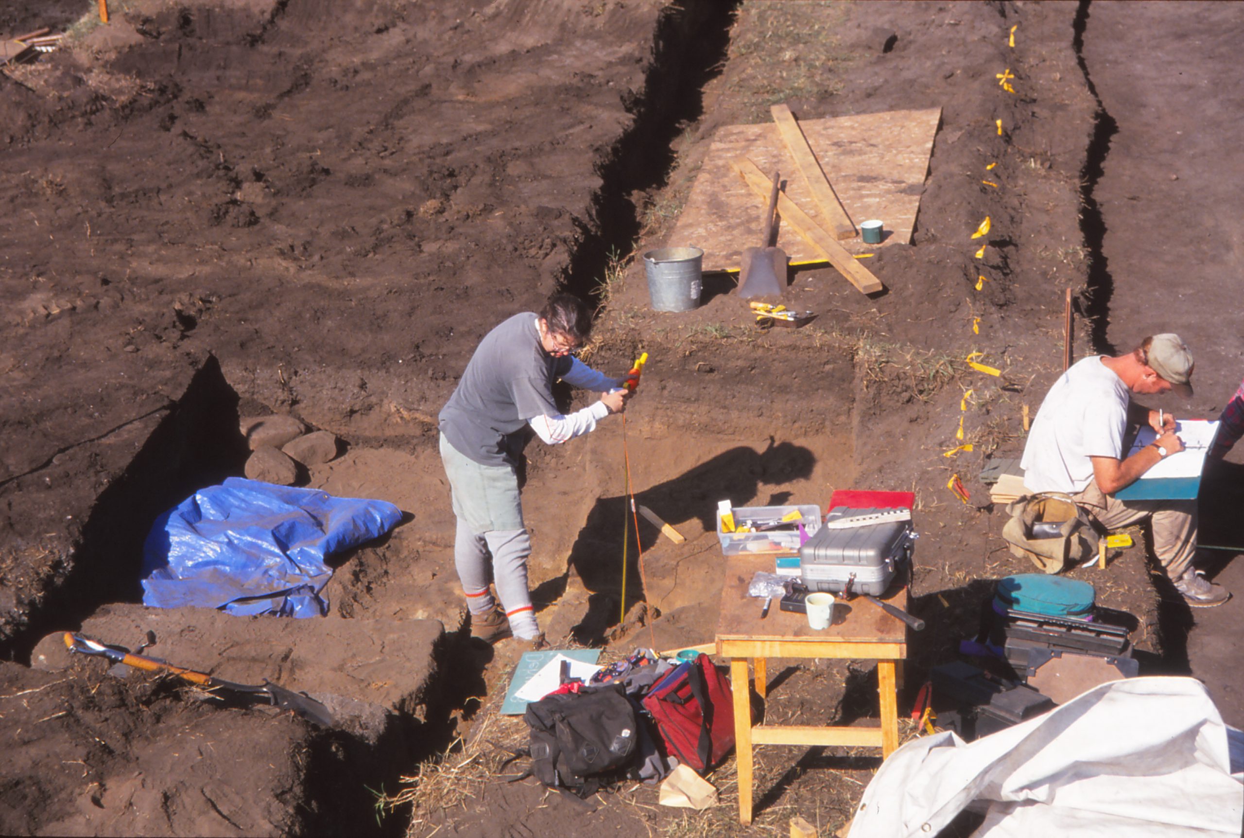 A photo copied from a 1994 slide shows archaeologists looking over tables with equipment surrounded by dirt at an excavation site.