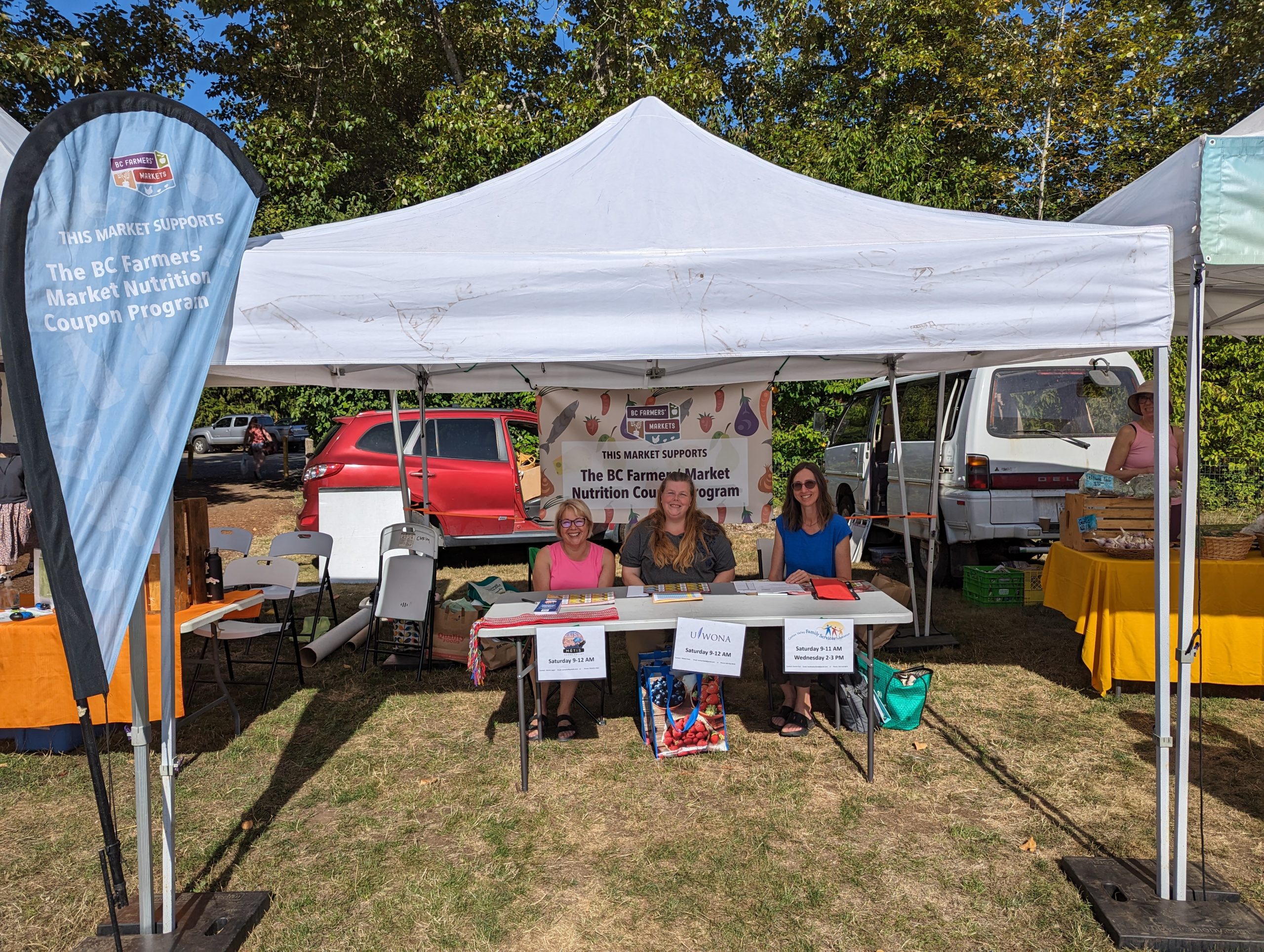 A table with three women sitting outside under a tent at the Comox Valley farmers' market