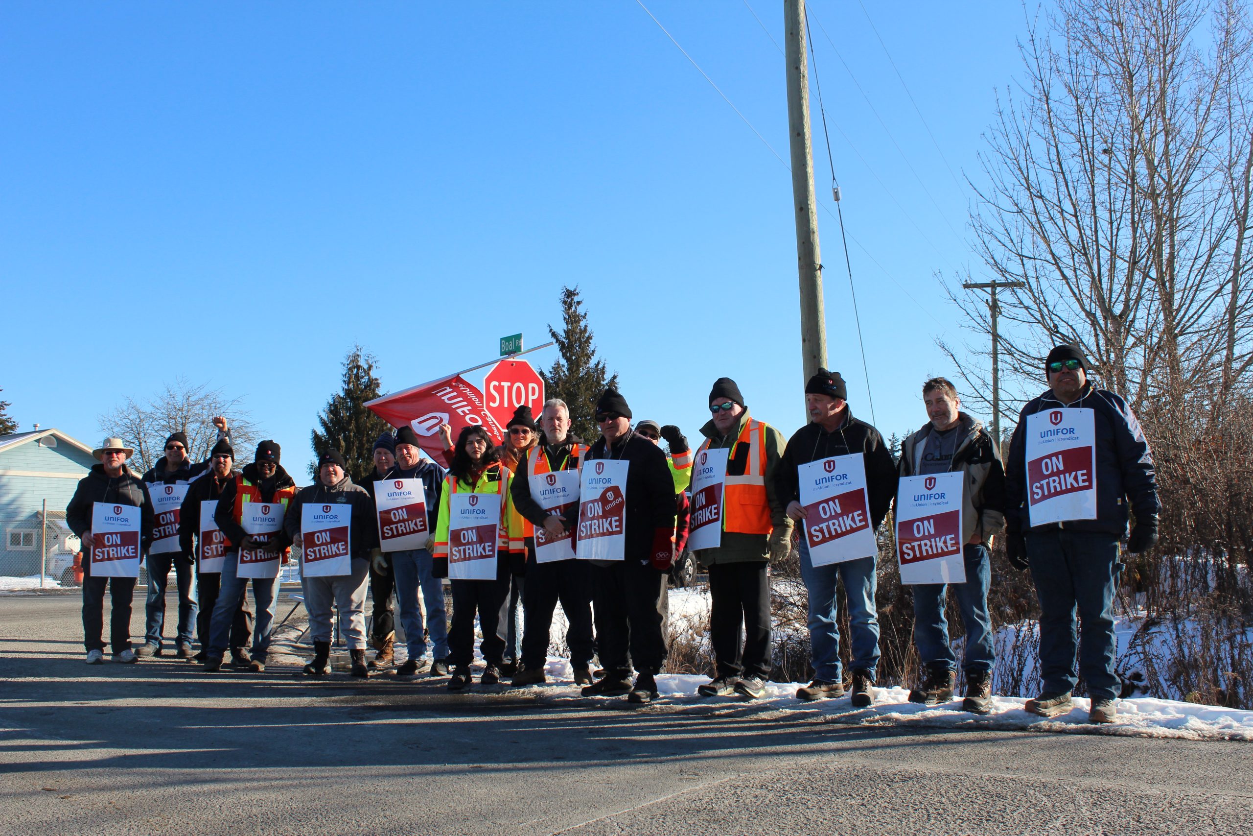 Picketers wearing signs that say "ON STRIKE" stand in a line along the road, some with fists in the air. They are part of the Cowichan Valley transit strike.