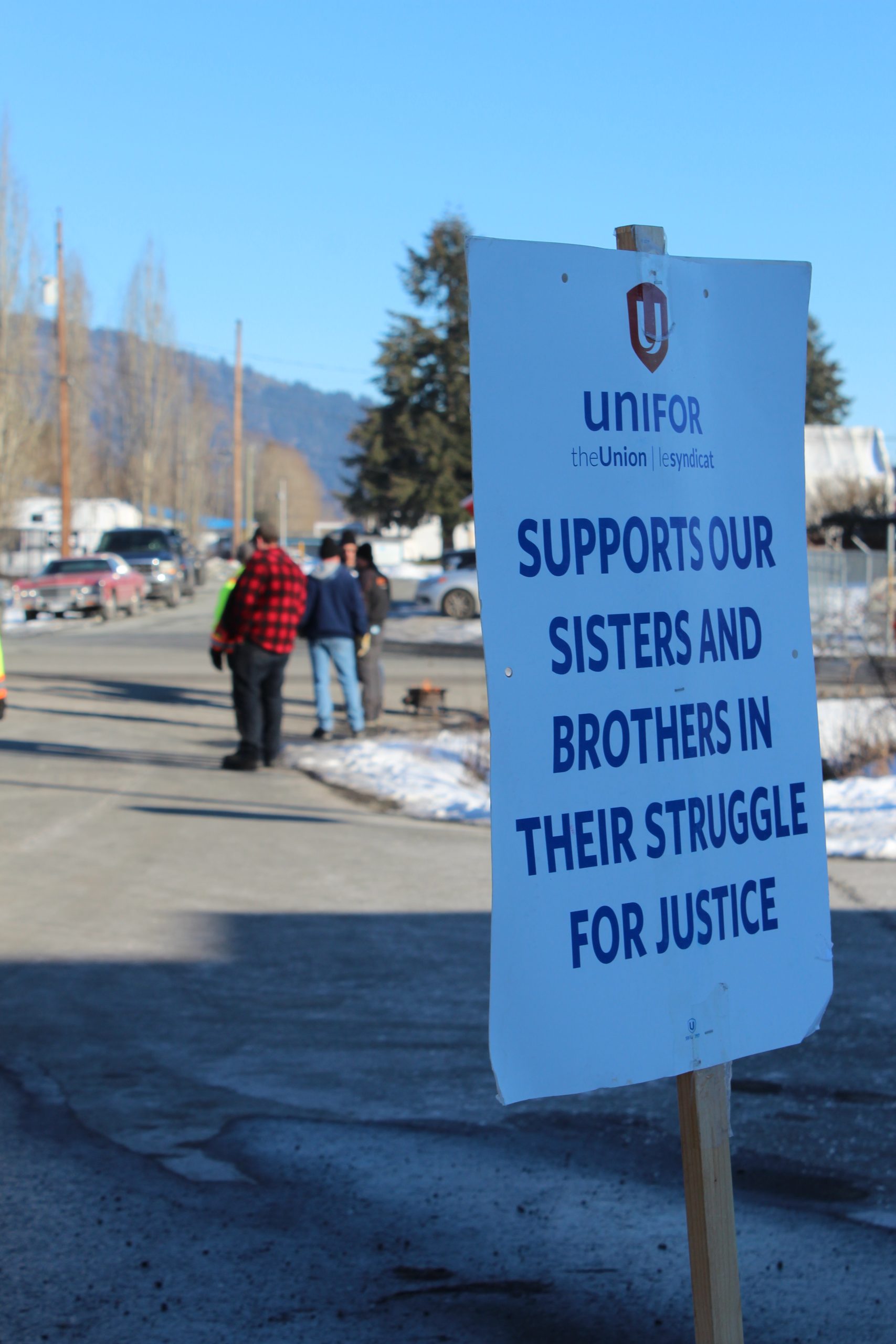 A Unifor sign that says "Supports our sisters and brothers in their struggle for justice." In the background are transit workers on strike.
