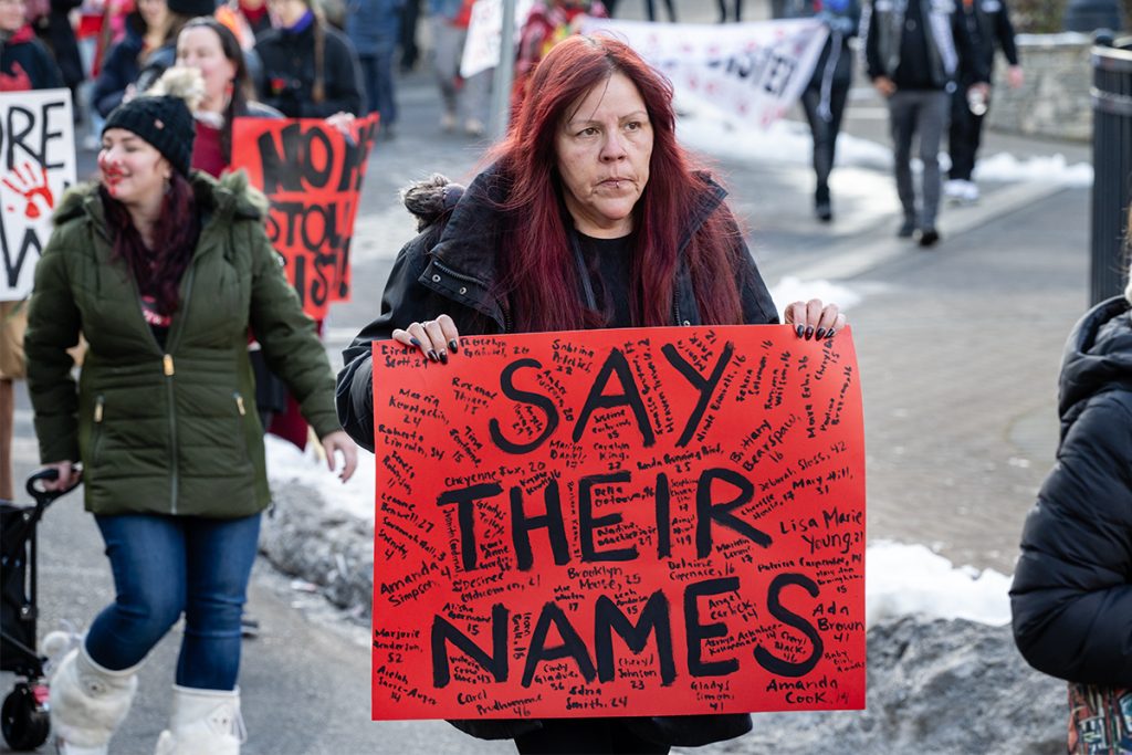 A woman carrying a sign reading "Say their names" with dozens of names in small print marches in downtown Nanaimo for missing and murdered Indigenous women and girls MMIWG2S+.