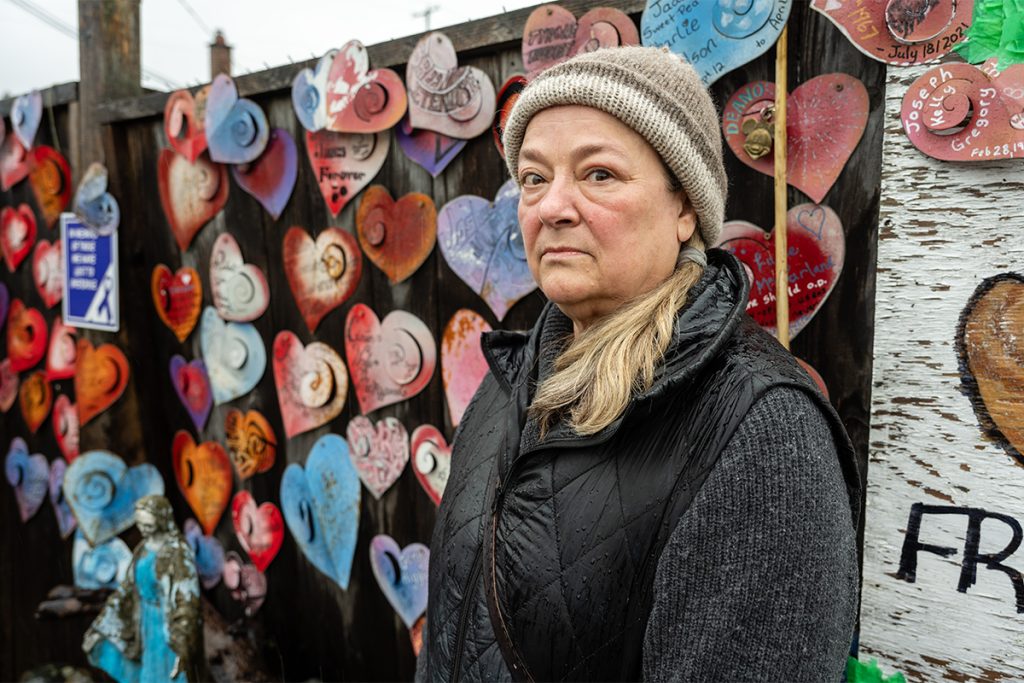 Photo of a woman standing in front of a fence with metal hearts bearing the names of people who died from overdoses.