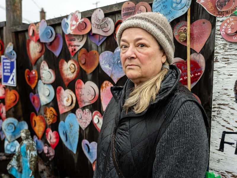 Photo of a woman standing in front of a fence with metal hearts bearing the names of people who died from overdoses.